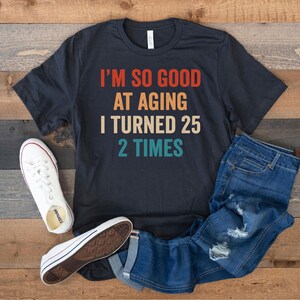 May include: A black t-shirt with a vintage-style graphic that reads "I'm so good at aging I turned 25 2 times". The shirt is laid out on a wooden surface with a pair of white sneakers and blue jeans.