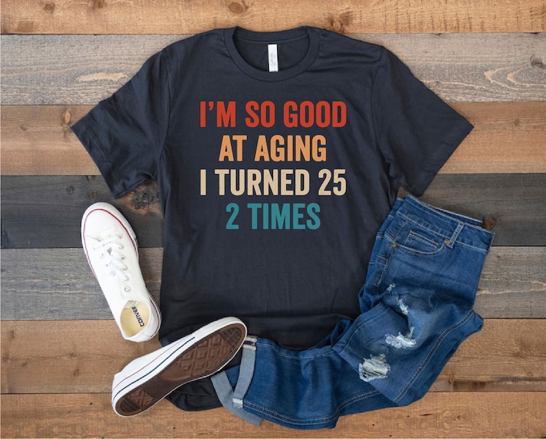 May include: A black t-shirt with a vintage-style graphic that reads "I'm so good at aging I turned 25 2 times". The shirt is laid out on a wooden surface with a pair of white sneakers and blue jeans.
