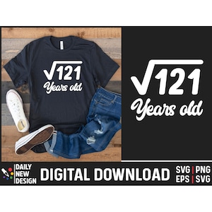 May include: A black t-shirt with a white graphic that says "√121 Years old". The t-shirt is laid out on a wooden surface with a pair of blue jeans and white sneakers.