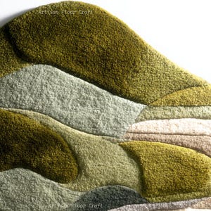 May include: A textured, abstract rug with a layered design in shades of green, gray, and beige. The rug features a soft, plush texture and a unique, organic shape. The words "Artisan Floor Craft" are printed on the rug.
