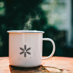 May include: White enamel mug with a black snowflake design and a blue rim. The mug is filled with a hot beverage and steam is rising from the top.