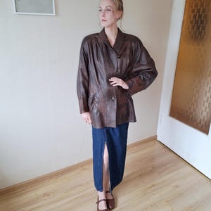 May include: A brown leather jacket with a button closure and a notched collar. The jacket is paired with a long denim skirt with a front slit. The outfit is completed with brown shoes.