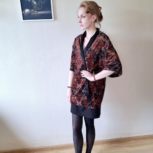 Könnte beinhalten: Gemusterter Kimono-Stil-Robe mit offenem Frontbereich in Rot-, Braun- und Schwarztönen. Die Robe hat eine schwarze Einfassung und wird über einem schwarzen Kleid getragen. Die Ärmel sind kurz und der Stoff hat ein Blumenmuster.