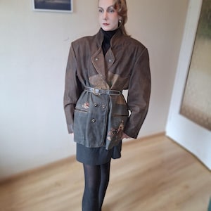 May include: A brown leather jacket with a unique design. The jacket features a double-breasted front, a belted waist, and decorative embroidery on the pockets. The jacket is worn over a black turtleneck and a dark skirt.