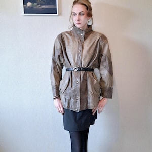 Può includere: Giacca vintage in pelle marrone con collo alto e chiusura con bottoni. La giacca presenta una cintura nera che stringe la vita e un design patchwork. La modella indossa una gonna e calze nere.