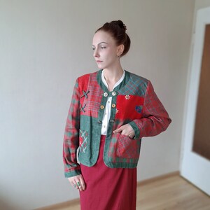 Könnte beinhalten: Eine Patchwork-Jacke mit rot-grünen Karopaneelen, verziert mit dekorativen Nähten und Holzknöpfen. Die Jacke hat einen runden Ausschnitt und zwei Vordertaschen. Das Model trägt ein weißes Hemd und einen roten Rock.