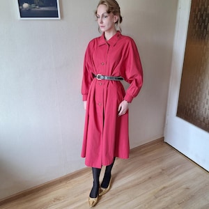 Può includere: Un lungo vestito rosso con bottoni sul davanti e una cintura nera che stringe la vita. L'abito ha maniche lunghe ed è abbinato a collant neri e scarpe color oro. L'abito è lungo fino al polpaccio.