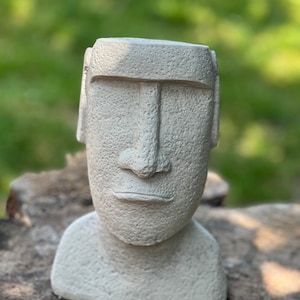 May include: A white concrete planter shaped like a bust of a Moai statue. The planter has a textured surface and a simple, minimalist design.
