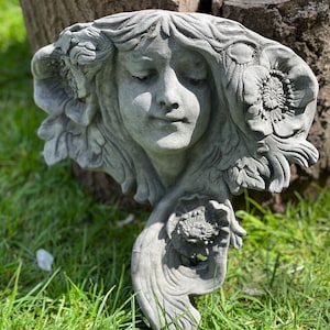 May include: A grey stone sculpture of a woman's face with flowers in her hair. The sculpture is in a garden setting.
