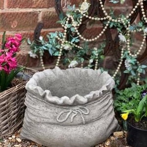 May include: A grey concrete planter shaped like a sack with a tied rope detail. The planter is sitting on a bed of gravel.