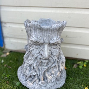 May include: A grey, weathered-effect planter sculpted to resemble a tree trunk with a carved face. The face features a long beard and leaf details. The planter is placed on a grassy surface.