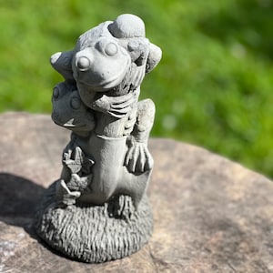 May include: A grey concrete garden statue of two frogs, one perched on top of the other, with a whimsical design.