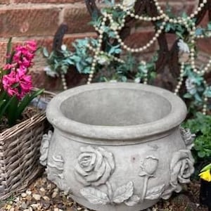 May include: A grey concrete planter with rose detailing. The round planter has a wide opening and is surrounded by decorative elements, including a wicker basket with pink flowers.