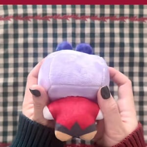 Puede incluir: Un peluche con forma de personaje, sostenido en las manos. El juguete tiene una cabeza de color lila claro con dos pompones azules, un cuerpo rojo y detalles en beige y negro. El fondo es un patr&oacute;n de cuadros verdes y blancos.
