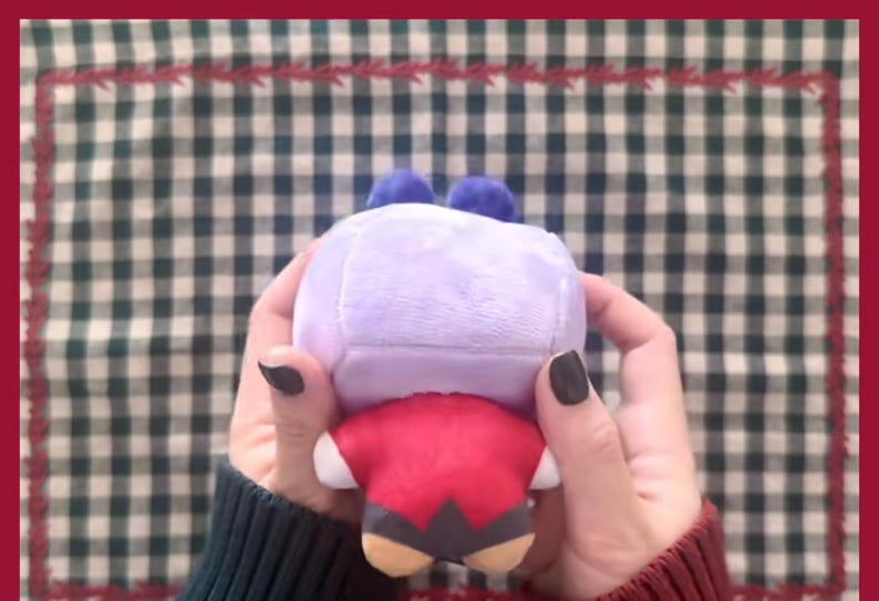 Puede incluir: Un peluche con forma de personaje, sostenido en las manos. El juguete tiene una cabeza de color lila claro con dos pompones azules, un cuerpo rojo y detalles en beige y negro. El fondo es un patr&oacute;n de cuadros verdes y blancos.