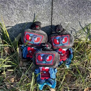 May include: Three plush toys with a robot design. Each has a black hat, a grey body with a screen-like face featuring red eyes, and a red bow tie. The toys are wearing blue jackets with red and white striped shirts.