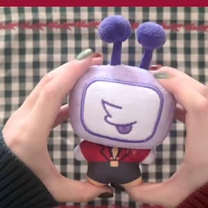 Puede incluir: Un peluche con forma de televisor retro, sostenido por manos. El televisor tiene un cuerpo morado, una pantalla blanca con una cara dibujada y dos protuberancias en forma de antena. Lleva una chaqueta roja y pantalones cortos negros.