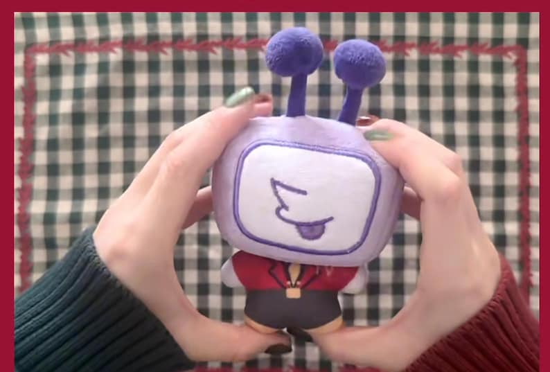 Puede incluir: Un peluche con forma de televisor retro, sostenido por manos. El televisor tiene un cuerpo morado, una pantalla blanca con una cara dibujada y dos protuberancias en forma de antena. Lleva una chaqueta roja y pantalones cortos negros.