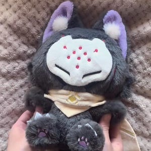 以下が含まれることがあります： グレーの毛皮、紫の耳、赤い斑点のある白い顔を持つ、様式化された生き物のぬいぐるみ。ベージュのスカーフと紫色の肉球が付いた爪のある足があります。柔らかく抱きしめたくなるおもちゃです。