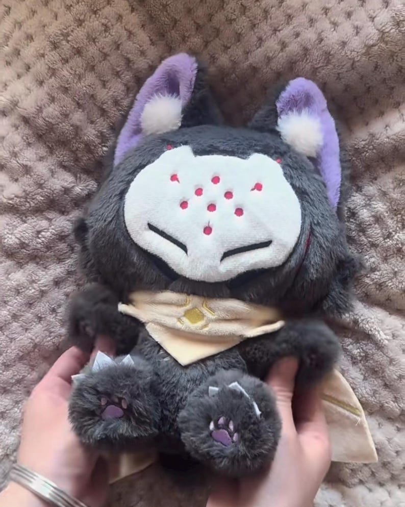 May include: A plush toy featuring a stylized creature with gray fur, purple ears, and a white face with red spots. It has a tan scarf and clawed feet with purple paw pads. The toy is soft and cuddly.