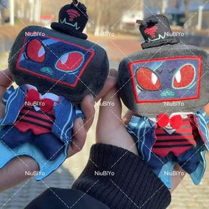 May include: Two plush toys resembling old televisions, each with a black body, red-eyed face, and a small hat with a wifi symbol. They wear blue and red outfits with bow ties. The toys are held in someone's hands.