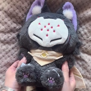 May include: A plush toy wolf with gray fur, purple ears, and a white face with red spots. It has a cream-colored scarf with a yellow emblem and purple paw pads. The toy is held by hands with a silver bracelet.