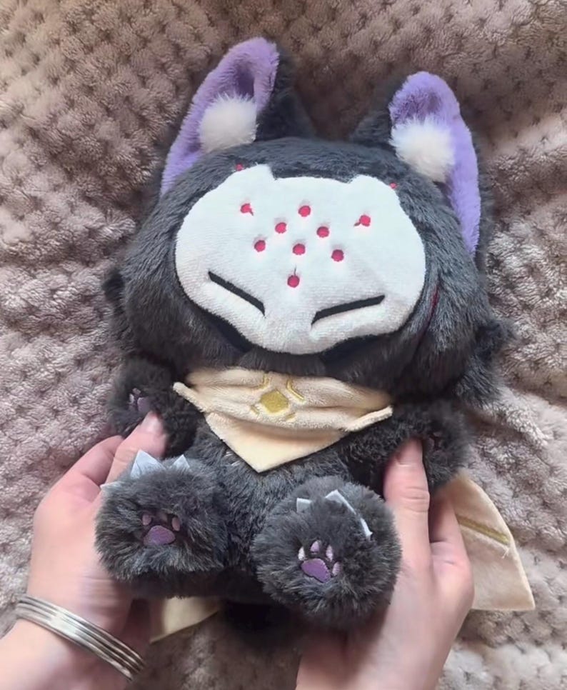May include: A plush toy wolf with gray fur, purple ears, and a white face with red spots. It has a cream-colored scarf with a yellow emblem and purple paw pads. The toy is held by hands with a silver bracelet.