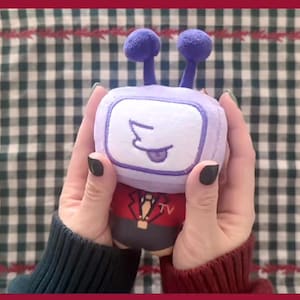 Puede incluir: Un peluche con forma de televisor retro, sostenido con dos manos. El televisor tiene un marco y antenas moradas, una pantalla blanca con una cara de dibujos animados y un traje rojo y negro. El fondo es un patr&oacute;n de cuadros verdes y blancos.