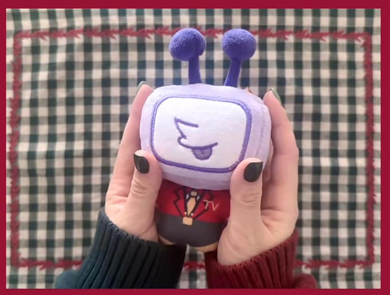 Puede incluir: Un peluche con forma de televisor retro, sostenido con dos manos. El televisor tiene un marco y antenas moradas, una pantalla blanca con una cara de dibujos animados y un traje rojo y negro. El fondo es un patr&oacute;n de cuadros verdes y blancos.