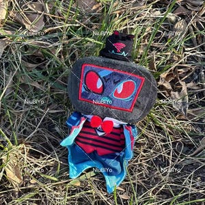 May include: A plush toy with a dark grey head, red-eyed face in a red-bordered square, and a black hat with a red emblem. It has a red and blue outfit with a heart-shaped detail. The toy is on grass and leaves.