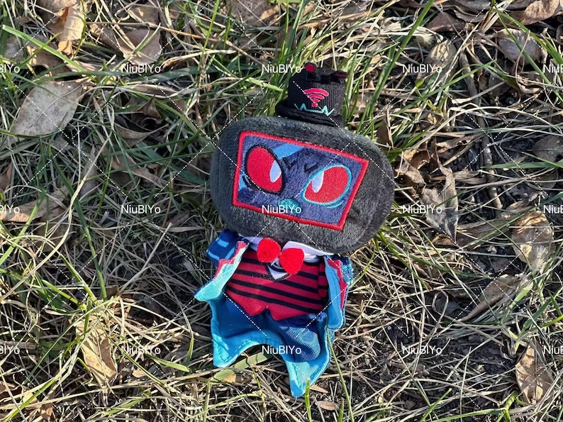 May include: A plush toy with a dark grey head, red-eyed face in a red-bordered square, and a black hat with a red emblem. It has a red and blue outfit with a heart-shaped detail. The toy is on grass and leaves.