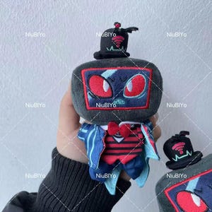 May include: A plush toy in the shape of a retro television set, complete with a top hat. The toy is grey with a screen showing red eyes and a blue design. It wears a red bow tie and a blue and red outfit. Another toy is also visible.