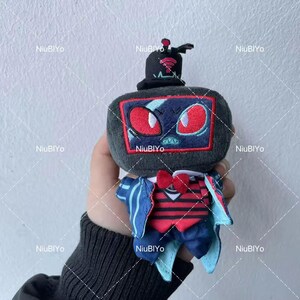 May include: A plush toy in the shape of a television set, complete with a top hat. The screen features red eyes and a blue design. It has a red bow tie and a striped vest, with blue and white accents.