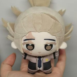 May include: A plush doll with a light tan head and spiky hair. The doll has large, expressive eyes and a small mouth. It wears a jacket with a blue tie and black pants. The doll is held in a hand.