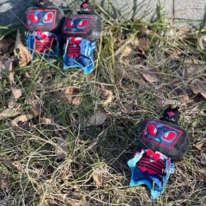 May include: Several plush robot-like figures with black bodies, red and blue accents, and antenna-like features are scattered on grass. The robots have rectangular faces with red eye designs and are wearing blue and red outfits.