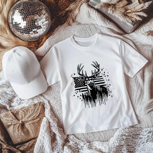 May include: White t-shirt with a black and white graphic of a deer with antlers standing in front of an American flag. The graphic is surrounded by stars.