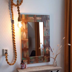 Pode incluir: Espelho rústico de parede com moldura de madeira composta por tábuas de várias cores. Uma luminária decorativa de corda pende da parede, iluminando uma pequena prateleira com um vaso e artigos de higiene pessoal. O ambiente é acolhedor.