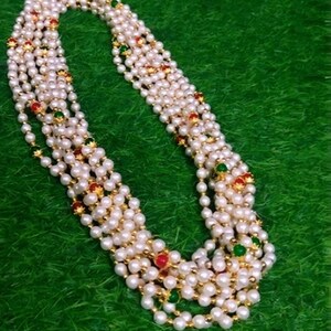 Puede incluir: Un collar de perlas de múltiples hilos con detalles rojos y verdes. El collar presenta pequeñas perlas redondas ensartadas con espaciadores dorados. El collar se muestra sobre una superficie verde.