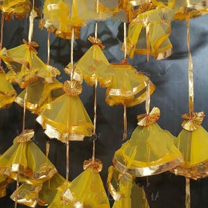 May include: A collection of hanging, yellow, cone-shaped decorations with gold trim and accents. The decorations are suspended from thin, gold-colored strings, creating a layered effect. The background is a dark, textured surface.
