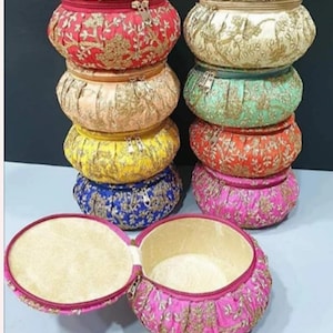 Könnte beinhalten: Eine Sammlung farbenfroher, runder Stoff-Aufbewahrungsbehälter mit goldener Stickerei. Die Behälter sind in zwei Säulen gestapelt, wobei einer geöffnet ist, um einen beigen Innenraum zu zeigen. Farben sind Rot, Gold, Gelb, Blau, Pink, Orange und Türkis.