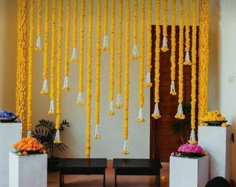 May include: A yellow flower garland backdrop with white accents, used for a wedding ceremony or other special event. The garland is hung on a wall and creates a beautiful and festive atmosphere.