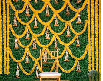 May include: A gold chair sits in front of a green wall decorated with yellow flower garlands. The wall is covered in green foliage and the garlands are arranged in a cascading pattern. The chair is placed in front of a round, gold-coloured platform filled with yellow flower petals.