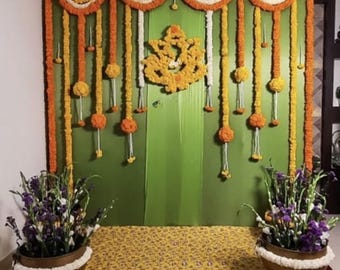 May include: A colourful backdrop for a celebration, with a green fabric wall decorated with marigold garlands in orange, yellow, and white. A floral Ganesh design is central. Two bronze pots with purple and white flowers are at the front.
