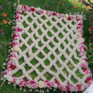 May include: A square, decorative floral arrangement with a lattice design. The lattice is made of white, fluffy material, and the edges are adorned with pink, red, and white roses. The arrangement is set on a bed of green grass.