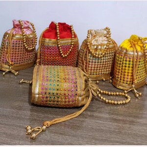 Puede incluir: Cinco bolsas decorativas con cordón en varios colores, incluyendo rosa, rojo, amarillo y dorado. Cada bolsa presenta intrincados abalorios y adornos dorados, con un cierre de cordón y detalles de borlas. Las bolsas están dispuestas sobre una superficie de madera.
