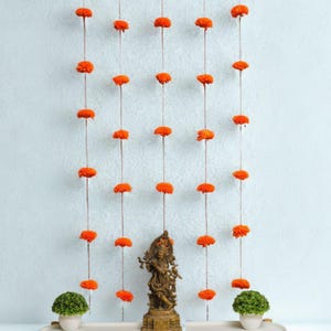 May include: Orange marigold flower garlands hanging against a light blue wall. A bronze statue of a deity and two small potted green plants sit on a white tray below the garlands. The flowers are evenly spaced.