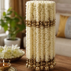 May include: A cylindrical floral garland made of cream-colored jasmine flowers, featuring horizontal bands of metallic gold beads and decorative gold-toned spherical pendants hanging from the bottom edge of the structure.