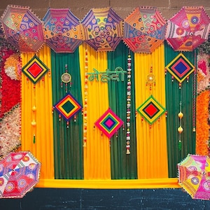 May include: A decorative backdrop featuring alternating vertical yellow and green fabric panels, adorned with five colorful patterned umbrellas at the top, two at the bottom, diamond-shaped wall hangings, and the Hindi text "मेहंदी" in green.