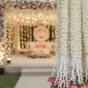 May include: Close-up of white floral garlands with pink accents, likely for a wedding or event. The garlands hang in front of a blurred background of a stage decorated with lights and flowers, creating a festive atmosphere.