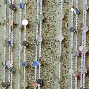 May include: Decorative beaded curtain featuring strands of clear beads and iridescent, round sequins. The sequins reflect various colors, including silver, blue, and pink. The curtain is designed to hang, adding a touch of sparkle and texture to any space.